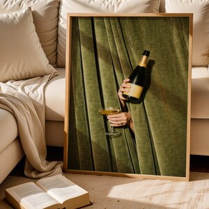 May include: A framed wall art print featuring a dark green velvet curtain with vertical folds. A hand emerges from behind the curtain, holding a dark glass bottle with a tan label and a stemmed glass filled with a golden liquid, creating a contrast.