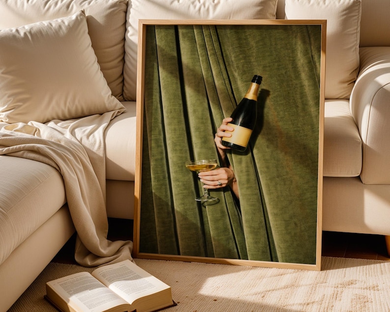 May include: A framed wall art print featuring a dark green velvet curtain with vertical folds. A hand emerges from behind the curtain, holding a dark glass bottle with a tan label and a stemmed glass filled with a golden liquid, creating a contrast.