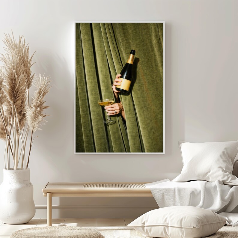 May include: A framed wall art print featuring a dark olive green velvet curtain backdrop. A hand emerges from the curtain holding a dark glass bottle with a gold label and a clear stemmed coupe glass filled with a golden liquid beverage.