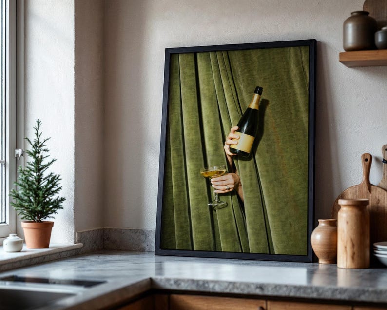 May include: A framed wall art print featuring a dark green velvet curtain backdrop. A bottle of champagne and a filled coupe glass are held through the fabric folds. The composition highlights the contrast between the deep green textile and the glass.
