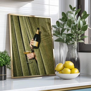 May include: A framed wall art print featuring a pair of hands emerging from olive green velvet curtains, holding a champagne bottle and a filled coupe glass. The artwork is displayed in a thin gold frame, showcasing a rich, textured fabric backdrop.