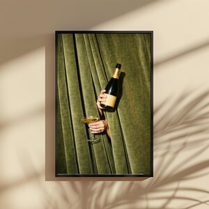 May include: A framed wall art print featuring a dark glass bottle with a tan label and a clear stemmed glass filled with a golden liquid, both held by hands emerging from behind heavy, pleated olive green velvet curtains.