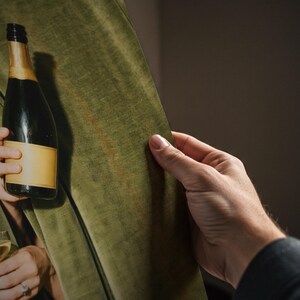 May include: A dark green velvet fabric drape featuring a subtle sheen and a smooth texture. A dark glass bottle with a blank, light-colored rectangular label is positioned against the fabric, showing a classic cylindrical shape and neck.