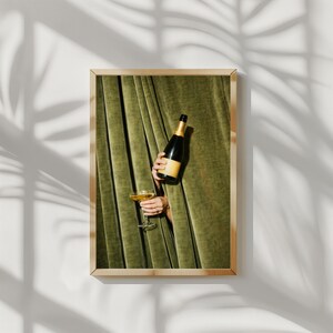 May include: A framed wall art print featuring a dark green velvet curtain backdrop. A hand emerges from the curtain holding a dark glass bottle with a gold foil neck and a clear stemmed glass filled with a golden liquid beverage.