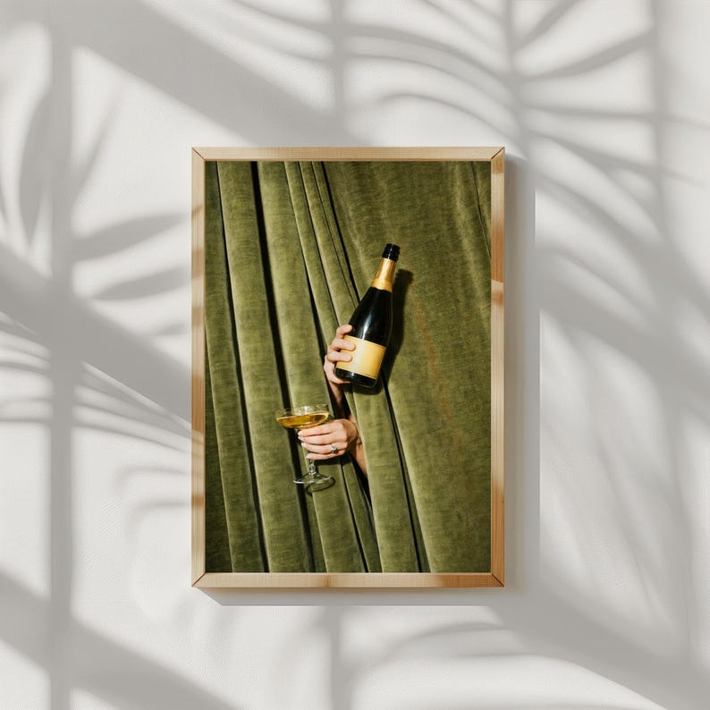 May include: A framed wall art print featuring a dark green velvet curtain backdrop. A hand emerges from the curtain holding a dark glass bottle with a gold foil neck and a clear stemmed glass filled with a golden liquid beverage.