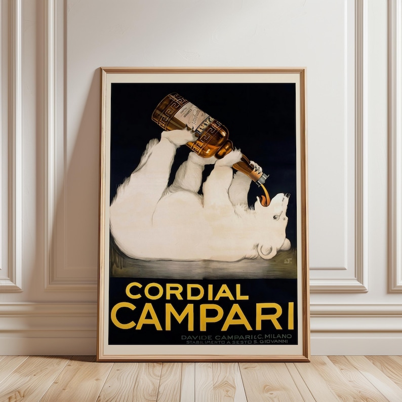 Pu&ograve; includere: Poster vintage incorniciato per Cordial Campari. L'opera presenta un orso polare bianco che beve da una bottiglia. Il poster ha uno sfondo nero con scritte dorate e una cornice in legno marrone.