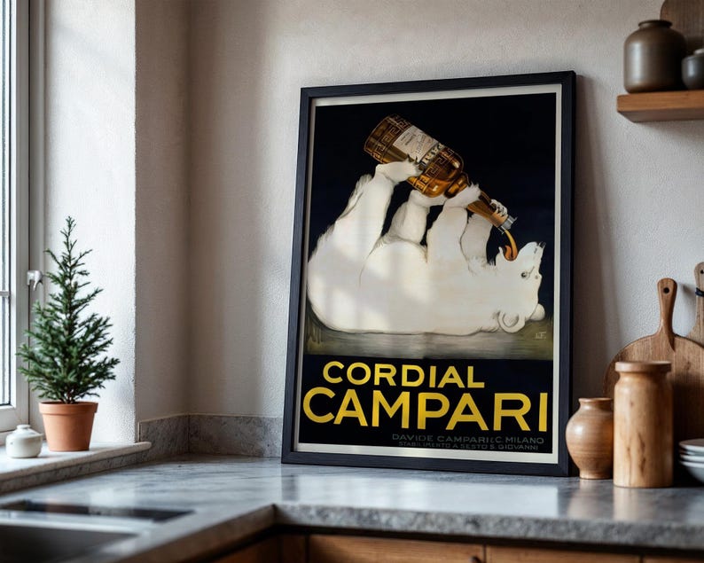 Pu&ograve; includere: Un poster vintage incorniciato che pubblicizza Cordial Campari. Raffigura un orso polare bianco che beve da una bottiglia di Campari. Il testo "Cordial Campari" &egrave; in lettere dorate su sfondo blu scuro.