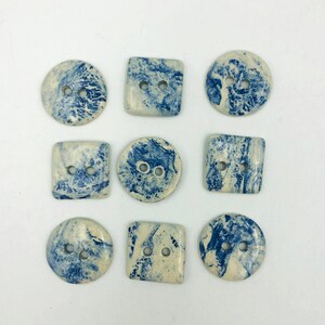 Pu&ograve; includere: Nove bottoni in ceramica di varie forme: rotondi e quadrati. Ogni bottone ha una base color crema con marmorizzazione blu e due fori per cucire. I bottoni sono disposti su una superficie bianca.