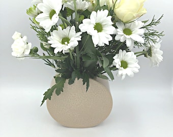 Pale pink ceramic windowsill vase with floral texture on one side and blue hydrangea bloom with green leaves on the other