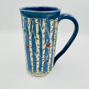 May include: A tall ceramic mug featuring a blue background with white birch tree trunks and small red birds. The mug has a solid dark blue rim and a matching curved handle. The design wraps around the cylindrical body, showcasing a winter forest scene.