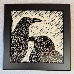 May include: A framed square art print featuring a black and white linocut style illustration of two ravens standing in the rain. The birds are depicted with detailed feather textures against a cream background with diagonal black rain lines.