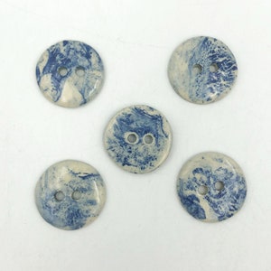 Pu&ograve; includere: Cinque bottoni rotondi con un design marmorizzato nei toni del blu e del crema. Ogni bottone ha due fori per cucire. I bottoni sono in ceramica.