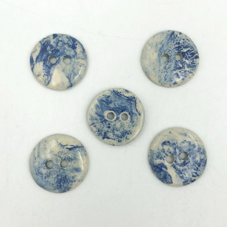 Pu&ograve; includere: Cinque bottoni rotondi con un design marmorizzato nei toni del blu e del crema. Ogni bottone ha due fori per cucire. I bottoni sono in ceramica.