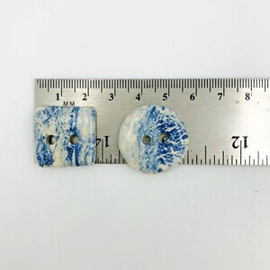 Pu&ograve; includere: Due bottoni in ceramica con un motivo marmorizzato blu e bianco. Uno &egrave; quadrato, l'altro rotondo. Ogni bottone ha due fori per cucire. I bottoni sono misurati con un righello in millimetri.