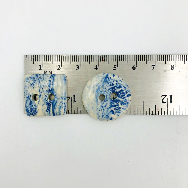 Pu&ograve; includere: Due bottoni in ceramica con un motivo marmorizzato blu e bianco. Uno &egrave; quadrato, l'altro rotondo. Ogni bottone ha due fori per cucire. I bottoni sono misurati con un righello in millimetri.