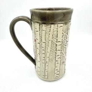 May include: A tall ceramic mug featuring a cream-colored surface with an embossed forest pattern of birch trees. The mug has a dark brown glazed rim and a matching curved handle. Small, subtle red accents are visible among the tree branches.