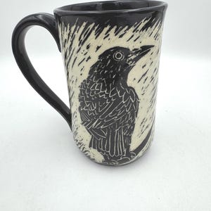 May include: A ceramic mug with a glossy black handle and rim, featuring a cream-colored body decorated with a black sgraffito-style illustration of a crow perched on a branch, surrounded by etched vertical lines that suggest a textured background.