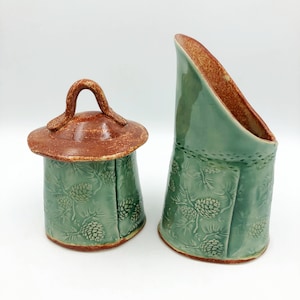 May include: A set of two ceramic containers with a green glaze and brown accents. The containers feature a pine cone design and are perfect for storing sugar and creamer.