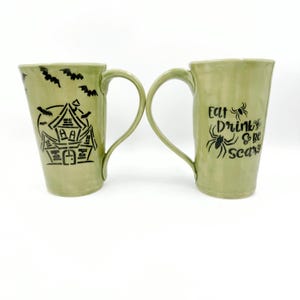 May include: Two light green ceramic mugs with black ink designs. The left mug features a haunted house and flying bats. The right mug displays the text Eat Drink & be scary with spider illustrations. Both mugs have a tall, tapered shape.