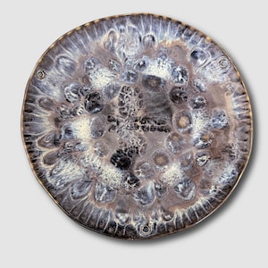 May include: A round ceramic plate with a unique, textured surface. The plate features a blend of colors, including shades of brown, gray, and white, creating a marbled effect. The rim has a metallic gold finish, and there are three small holes evenly spaced around the edge.