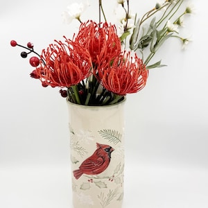 May include: A cylindrical cream-colored ceramic vase featuring a painted red cardinal bird, green holly leaves, and white snowflakes. The vase is filled with a bouquet of vibrant red pincushion protea flowers, small red berries, and white blossoms.