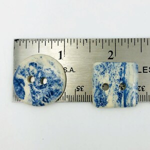 Pu&ograve; includere: Due bottoni decorativi, uno rotondo e uno quadrato, con un design marmorizzato blu e bianco. Il bottone rotondo ha due fori e misura circa 2,5 cm di diametro. Anche il bottone quadrato ha due fori.