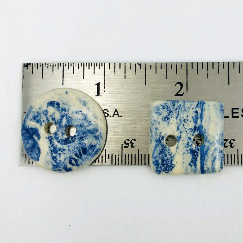 Pu&ograve; includere: Due bottoni decorativi, uno rotondo e uno quadrato, con un design marmorizzato blu e bianco. Il bottone rotondo ha due fori e misura circa 2,5 cm di diametro. Anche il bottone quadrato ha due fori.