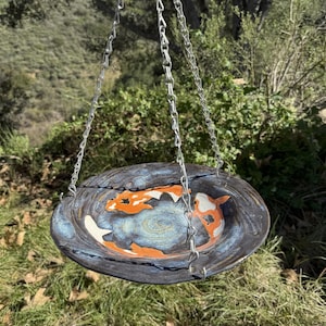 May include: A hanging ceramic bird bath with a blue and black glazed finish. The bird bath features a koi fish design in orange and white. The bird bath is suspended by silver chains and is set against a backdrop of green foliage.