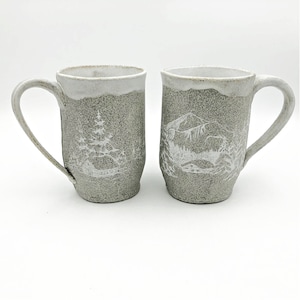 May include: Two ceramic mugs with white glaze and a speckled gray finish. One mug features a white silhouette of a mountain range, while the other mug features a white silhouette of a forest.