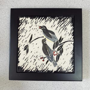 May include: A framed square art piece featuring two stylized birds perched on a branch with berries. The birds are depicted in shades of gray and black against a white background with black diagonal lines. The frame is black.