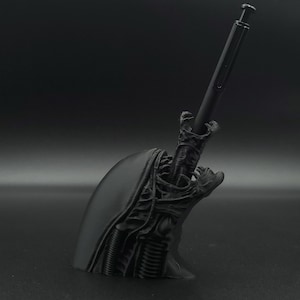 May include: Black 3D printed alien head pen holder with a black pen sticking out of the top of the head.