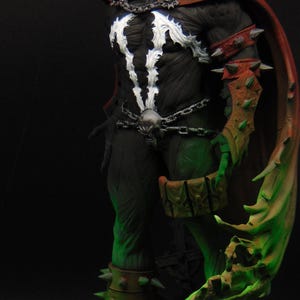 May include: A Spawn action figure featuring a black suit with a white chest emblem, a flowing red cape, and silver chain details. The character wears spiked arm and leg guards, a skull-adorned belt, and stands on a base with green sculpted details.