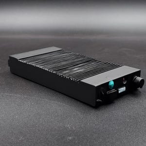 May include: A black rectangular electronic device with a ridged top and a small, round knob on one end. The device has a small, turquoise-colored light and several ports. The device is approximately 6 inches long.
