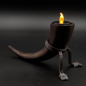 May include: A dark brown, horn-shaped candle holder with a flickering LED flame. The holder is supported by a metal stand with a circular band and two feet. The candle holder is designed to resemble a drinking horn.