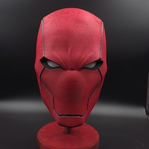 May include: A red superhero mask with a textured, mesh-like surface. The mask has black eye openings and a defined facial structure. It is displayed on a red stand against a dark background, highlighting the intricate design.