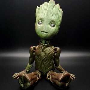 May include: A green and brown figurine of a character from the Guardians of the Galaxy movie franchise. The character is sitting with its legs crossed and has a serious expression on its face.