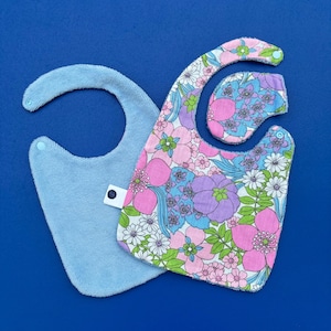 Könnte beinhalten: Drei Baby-Lätzchen auf blauem Hintergrund. Eines ist aus hellblauem Frottee. Die anderen beiden haben ein Blumenmuster mit rosa, lila, blauen und weißen Blumen. Die Lätzchen sind so konzipiert, dass sie Spritzer und Sabber auffangen.