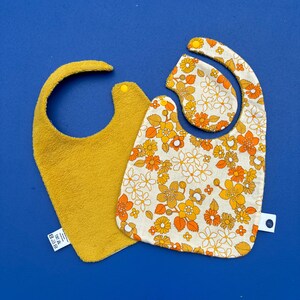 Könnte beinhalten: Zwei Baby-Lätzchen, eines aus gelbem Frottee und eines weiß mit einem Retro-Blumenmuster in Orange und Gelb.