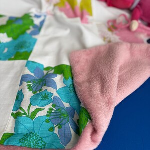 Peut inclure: Une courtepointe patchwork avec un motif floral dans les tons de bleu, vert et blanc. La courtepointe est bordée d'un tissu en peluche rose doux. Une poupée rose est partiellement visible en arrière-plan.