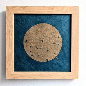Op de afbeelding: Een ingelijst kunstwerk met een blauwe achtergrond en een grote, ronde, gouden cirkel in het midden. De cirkel is gevuld met kleine, donkerblauwe vogelsilhouetten.
