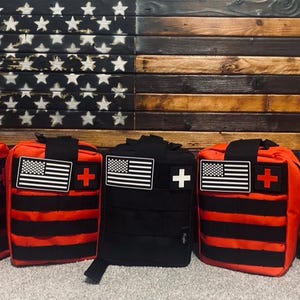 Puede incluir: Cinco bolsas de primeros auxilios rojas y negras con un parche de bandera estadounidense blanco y negro y un parche de cruz roja en cada bolsa. Las bolsas están alineadas en una fila frente a un fondo de madera con una bandera estadounidense.