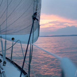 May include: A sailboat with a white sail is sailing on a body of water at sunset. The water is a light blue color and the sky is a light pink color.