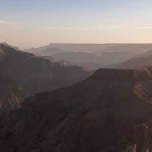 May include: A scenic landscape of a canyon at sunset. The image features layers of rock formations in shades of brown and gray, with a soft, hazy atmosphere. The sky transitions from blue to a warm, golden hue.