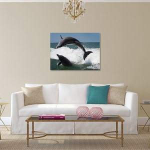 May include: Two dolphins leap out of the water, creating a spray of white foam. The dolphins are dark gray and are silhouetted against a blue sky and ocean.