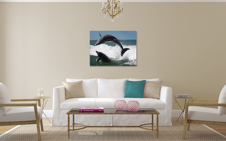 May include: Two dolphins leap out of the water, creating a spray of white foam. The dolphins are dark gray and are silhouetted against a blue sky and ocean.