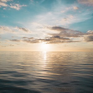 May include: A panoramic view of a calm ocean with a bright, golden sunset reflecting on the water. The sky is a mix of blue and white clouds.