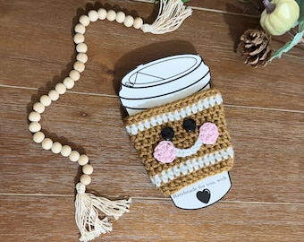 May include: A brown and white crocheted coffee cup cozy with a smiling face and pink cheeks. The cozy is handmade and has the text "Handmade for you, with" on the bottom.