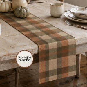 May include: A rectangular table runner featuring a plaid pattern in shades of muted orange, olive green, and beige. A circular white badge with a brown border is overlaid on the runner, displaying the black text 5 designs available.