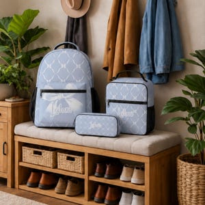 May include: A light blue backpack, lunch bag, and pencil case set featuring a white lattice pattern and a large white bow graphic on the backpack. Each item is personalized with the name Leilani in white script text and has black zippers.
