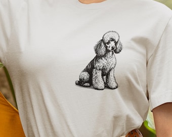 May include: A light-colored t-shirt featuring a detailed black and white illustration of a poodle. The poodle is depicted in a seated position, with its fluffy fur and distinctive features clearly visible. The shirt is a neutral color, providing a clean backdrop for the dog graphic.